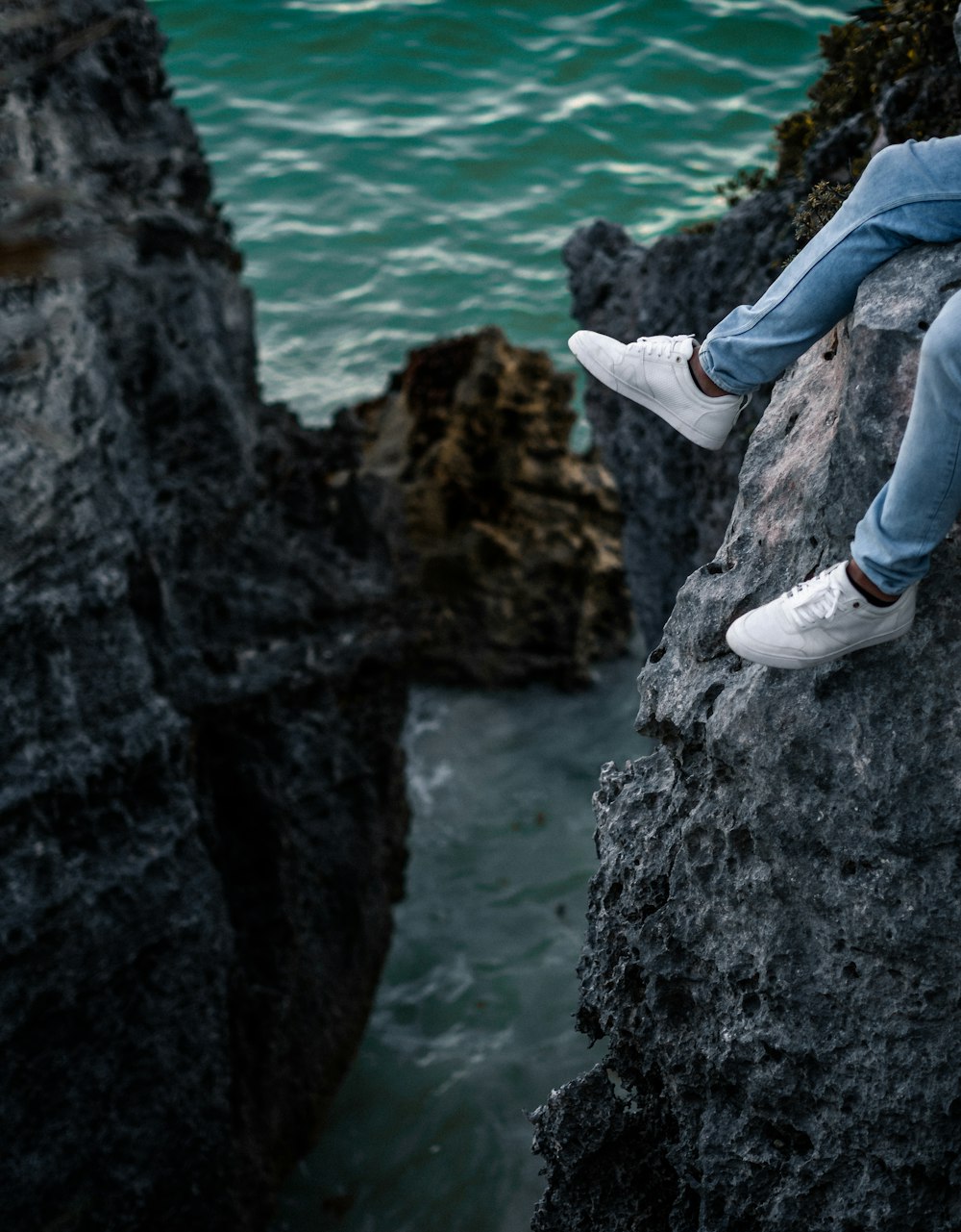 person in blue jeans and white sneakers sitting on rock cliff on sea