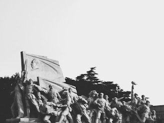 A large stone sculpture depicting a group of people, likely soldiers or workers, in dynamic poses. The monument has a prominent bas-relief of a face on a banner-like structure. Trees are visible in the background.