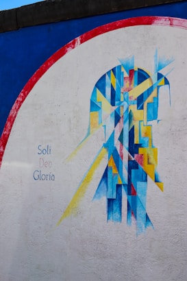 An abstract mural painted on a wall, featuring geometric shapes in blue, yellow, and red against a white background. The design resembles a stylized figure or object, with sharp angles and lines creating a dynamic composition. On the left side, the words 'Soli Deo Gloria' are written in a simple, elegant font.