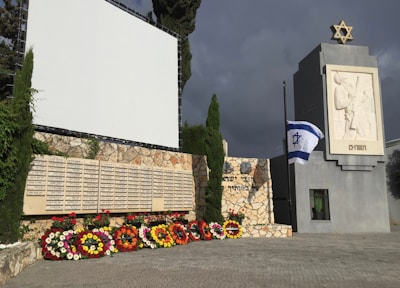 A memorial structure with Hebrew inscriptions, decorated with an array of colorful wreaths composed of various flowers. A prominent white and blue flag with a Star of David is flown at half-mast beside the structure, which features a large vertical monument engraved with soldier-like figures and a Star of David atop it. The backdrop includes a large blank screen and green trees under a cloudy sky.