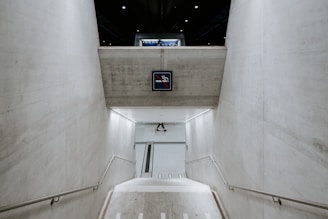 A modern, concrete staircase leads down into what appears to be an underground tunnel or passageway. The walls are stark and minimalistic, and a no smoking sign is prominently displayed above the stairs. Subtle lighting fixtures line the stairs, providing a soft illumination.