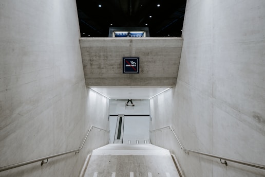 A modern, concrete staircase leads down into what appears to be an underground tunnel or passageway. The walls are stark and minimalistic, and a no smoking sign is prominently displayed above the stairs. Subtle lighting fixtures line the stairs, providing a soft illumination.
