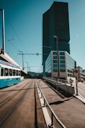 A modern urban setting featuring a large, dark glass skyscraper on the right side, with a tram moving along tracks in the foreground. The skyline is clear with minimal cloud presence, under a blue sky. The street is wide and well-paved, with some road signs visible.