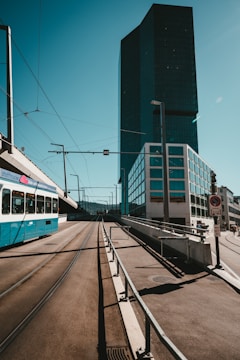 A modern urban setting featuring a large, dark glass skyscraper on the right side, with a tram moving along tracks in the foreground. The skyline is clear with minimal cloud presence, under a blue sky. The street is wide and well-paved, with some road signs visible.
