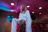 A person dressed in a white outfit is sitting on a chair in a dimly lit room. The space has a modern, industrial feel with exposed ceiling lights and colorful lighting casting blue and pink hues. Potted cacti and various plants are arranged on shelves in the background, and red and wooden chairs can be seen.