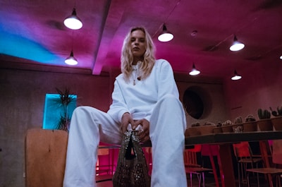 A person dressed in a white outfit is sitting on a chair in a dimly lit room. The space has a modern, industrial feel with exposed ceiling lights and colorful lighting casting blue and pink hues. Potted cacti and various plants are arranged on shelves in the background, and red and wooden chairs can be seen.