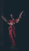 A dark and somber statue of an angel holding a scroll with the inscription 'GLORIA IN EXCELSIS DEO' draped across its outstretched arms. The angel is dressed in a flowing red robe and is partially illuminated against a dark background, enhancing the dramatic effect.