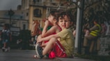 Two young children with curly hair are sitting on a pavement edge. The child in the foreground is wearing a yellow shirt and red shorts with a slightly tense expression. The other child is wearing a red dress and holding a brown paper bag. Surrounding them is an urban setting with buildings and blurred figures in the background, including a person wearing a fluorescent vest.
