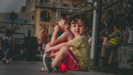 Two young children with curly hair are sitting on a pavement edge. The child in the foreground is wearing a yellow shirt and red shorts with a slightly tense expression. The other child is wearing a red dress and holding a brown paper bag. Surrounding them is an urban setting with buildings and blurred figures in the background, including a person wearing a fluorescent vest.