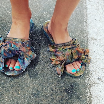 A close-up view of feet wearing colorful sandals with large, fabric bows. The toenails are painted in a bright turquoise color, and the ground appears to be textured asphalt.