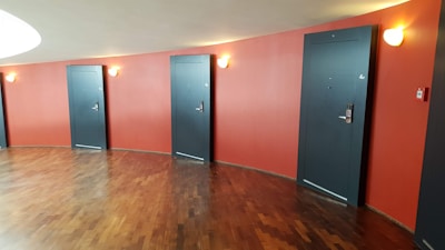 A hallway features several gray doors with room numbers, set against a curved red wall. The floor is made of polished wood, and wall sconces provide soft lighting.