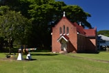 A small, red-brick church with a steeply pitched roof and a cross at the top stands in a grassy area. To the left, a multi-colored piece of machinery or artwork is situated on the lawn. The backdrop includes lush, green trees and clear blue skies.