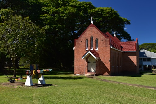A small, red-brick church with a steeply pitched roof and a cross at the top stands in a grassy area. To the left, a multi-colored piece of machinery or artwork is situated on the lawn. The backdrop includes lush, green trees and clear blue skies.