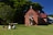 A small, red-brick church with a steeply pitched roof and a cross at the top stands in a grassy area. To the left, a multi-colored piece of machinery or artwork is situated on the lawn. The backdrop includes lush, green trees and clear blue skies.