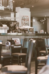 A cozy coffee shop interior with a modern espresso machine and cups arranged on the counter. Above the counter, there is a decorative sign that says 'CHANGE YOUR LIFE.' The room is filled with wooden chairs and round tables. A stylish decor includes a New York-themed picture and wine glasses on display.