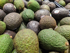 A pile of avocados with varying shades of green and brown skins, some with stickers. The texture of the avocados is bumpy and they are stacked closely together.