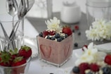A small glass container holds a creamy dessert topped with fresh strawberries, blueberries, and oats, garnished with a white flower. Additional strawberries are placed beside the glass, accompanied by a spoon and other utensils. A soft, neutral background includes more flowers and a glass of utensils, creating an elegant and inviting presentation.