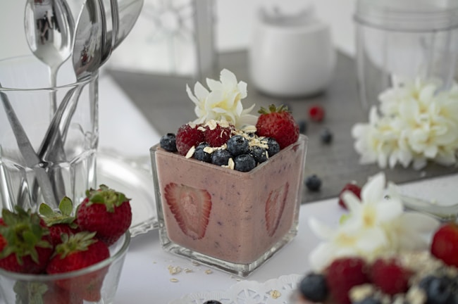 A small glass container holds a creamy dessert topped with fresh strawberries, blueberries, and oats, garnished with a white flower. Additional strawberries are placed beside the glass, accompanied by a spoon and other utensils. A soft, neutral background includes more flowers and a glass of utensils, creating an elegant and inviting presentation.