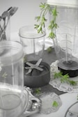 A collection of clear plastic blender containers and a blade attachment are arranged on a surface with decorative silverware and green plant sprigs scattered around. The scene includes doily-like placements, creating a mix of industrial and natural elements.