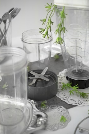 A collection of clear plastic blender containers and a blade attachment are arranged on a surface with decorative silverware and green plant sprigs scattered around. The scene includes doily-like placements, creating a mix of industrial and natural elements.