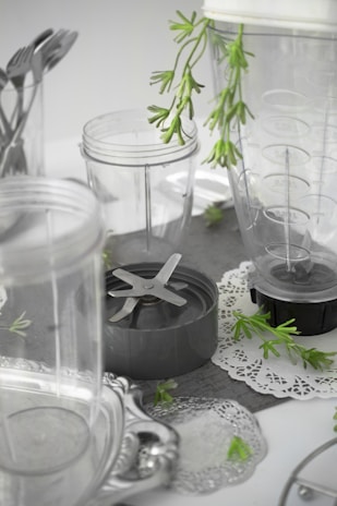 A collection of clear plastic blender containers and a blade attachment are arranged on a surface with decorative silverware and green plant sprigs scattered around. The scene includes doily-like placements, creating a mix of industrial and natural elements.