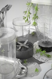 A collection of clear plastic blender containers and a blade attachment are arranged on a surface with decorative silverware and green plant sprigs scattered around. The scene includes doily-like placements, creating a mix of industrial and natural elements.
