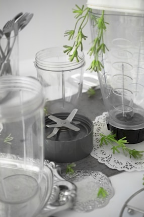 A collection of clear plastic blender containers and a blade attachment are arranged on a surface with decorative silverware and green plant sprigs scattered around. The scene includes doily-like placements, creating a mix of industrial and natural elements.