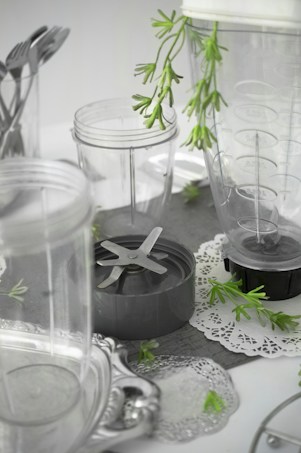 A collection of clear plastic blender containers and a blade attachment are arranged on a surface with decorative silverware and green plant sprigs scattered around. The scene includes doily-like placements, creating a mix of industrial and natural elements.