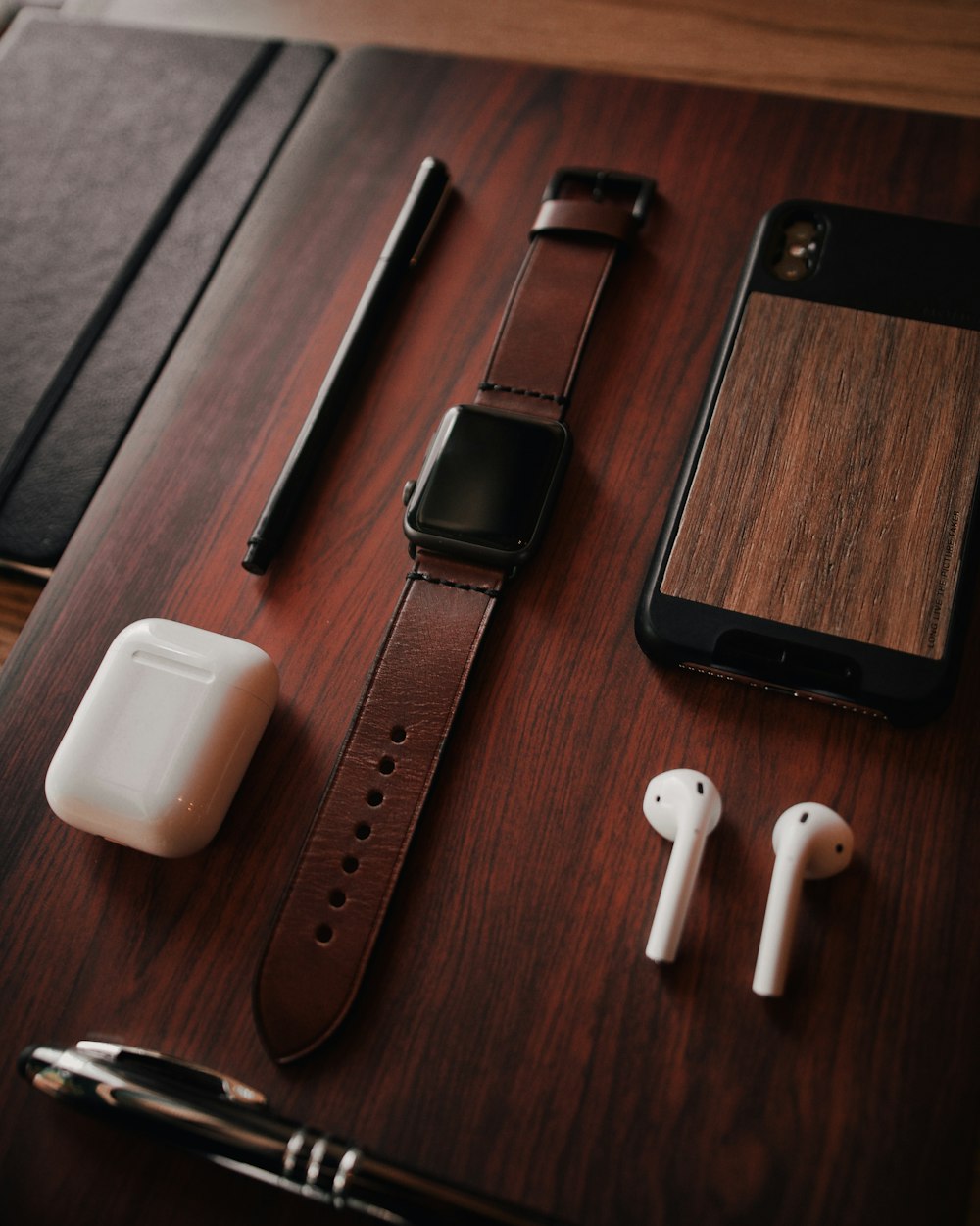 black aluminum Apple Watch with brown leather band, Apple AirPods with case, black pen, and brown and black case on brown wooden surface