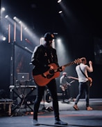 A band is performing on stage with a focus on a guitarist wearing a black cap and leather jacket. Stage lights illuminate the platform, creating a dramatic atmosphere. Other musicians in the background play instruments, including another guitar and electronic equipment.