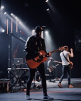 A band is performing on stage with a focus on a guitarist wearing a black cap and leather jacket. Stage lights illuminate the platform, creating a dramatic atmosphere. Other musicians in the background play instruments, including another guitar and electronic equipment.