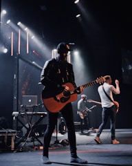 A band is performing on stage with a focus on a guitarist wearing a black cap and leather jacket. Stage lights illuminate the platform, creating a dramatic atmosphere. Other musicians in the background play instruments, including another guitar and electronic equipment.