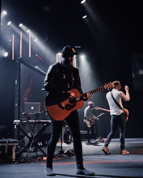 A band is performing on stage with a focus on a guitarist wearing a black cap and leather jacket. Stage lights illuminate the platform, creating a dramatic atmosphere. Other musicians in the background play instruments, including another guitar and electronic equipment.