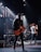 A band is performing on stage with a focus on a guitarist wearing a black cap and leather jacket. Stage lights illuminate the platform, creating a dramatic atmosphere. Other musicians in the background play instruments, including another guitar and electronic equipment.