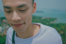 A close-up portrait of a person wearing a light-colored shirt. The background is blurred, depicting a vast landscape with greenery and a body of water, suggesting an outdoor setting. The person's expression is serene and thoughtful.
