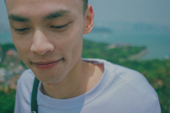 A close-up portrait of a person wearing a light-colored shirt. The background is blurred, depicting a vast landscape with greenery and a body of water, suggesting an outdoor setting. The person's expression is serene and thoughtful.