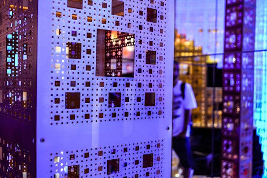 An intricate pattern of white perforated panels with square cutouts is prominently displayed, creating a layered and geometric effect. The background shows a blend of blue and purple lighting, adding a futuristic and abstract ambiance. A person is partially visible in the reflection, adding a sense of depth to the scene.