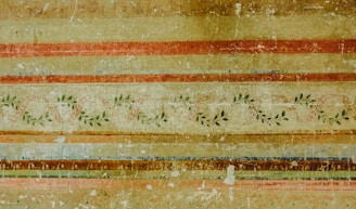 An ornate, vintage-looking wall mural featuring horizontal stripes in warm earth tones, including browns, reds, and golds. Interspersed among these stripes are intricate vine patterns with green leaves and winding ribbons, giving a classical and aged appearance.