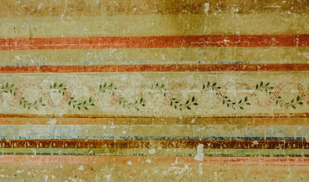 An ornate, vintage-looking wall mural featuring horizontal stripes in warm earth tones, including browns, reds, and golds. Interspersed among these stripes are intricate vine patterns with green leaves and winding ribbons, giving a classical and aged appearance.