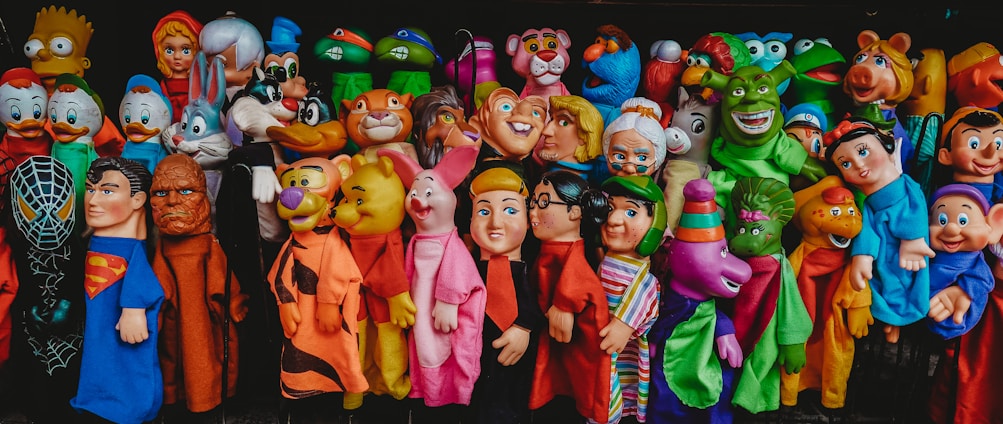A colorful assortment of character-themed puppets displayed in a vibrant array. Each puppet represents a distinct fictional character, featuring exaggerated facial features, diverse colors, and whimsical expressions, all lined up closely together.