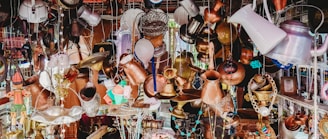 A chaotic array of assorted vintage and rustic metal objects hanging and displayed in an eclectic manner. The collection includes kettles, lamps, chains, and various other metallic containers, creating an antique and cluttered aesthetic.