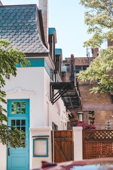 The image depicts a quaint urban courtyard area featuring a white building with distinct teal accents on the door and window frames. The architecture includes a roof with fish scale shingles and a charming wooden entry gate. A metal fire escape ladder adds an industrial touch while colorful flowers in a planter provide a splash of color against a wooden lattice fence. Nearby, a brick building rises in the background with windows suggestive of an older, urban setting. Trees add greenery to the scene, enhancing the cozy, inviting atmosphere.