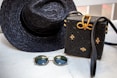 A woven black hat and a matching black woven handbag with gold accents are placed on a light wooden surface. Beside them, a pair of round sunglasses with dark lenses and thin golden frames rests. The woven handbag features a decorative bow made of golden material on top.