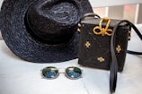 A woven black hat and a matching black woven handbag with gold accents are placed on a light wooden surface. Beside them, a pair of round sunglasses with dark lenses and thin golden frames rests. The woven handbag features a decorative bow made of golden material on top.