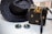 A woven black hat and a matching black woven handbag with gold accents are placed on a light wooden surface. Beside them, a pair of round sunglasses with dark lenses and thin golden frames rests. The woven handbag features a decorative bow made of golden material on top.