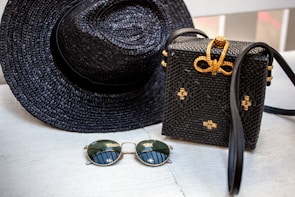 A woven black hat and a matching black woven handbag with gold accents are placed on a light wooden surface. Beside them, a pair of round sunglasses with dark lenses and thin golden frames rests. The woven handbag features a decorative bow made of golden material on top.