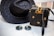 A woven black hat and a matching black woven handbag with gold accents are placed on a light wooden surface. Beside them, a pair of round sunglasses with dark lenses and thin golden frames rests. The woven handbag features a decorative bow made of golden material on top.