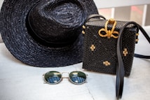 A woven black hat and a matching black woven handbag with gold accents are placed on a light wooden surface. Beside them, a pair of round sunglasses with dark lenses and thin golden frames rests. The woven handbag features a decorative bow made of golden material on top.