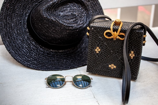A woven black hat and a matching black woven handbag with gold accents are placed on a light wooden surface. Beside them, a pair of round sunglasses with dark lenses and thin golden frames rests. The woven handbag features a decorative bow made of golden material on top.