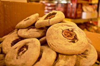 A close-up of several round, flat, grayish snacks, each with a dark, central filling, piled on top of one another inside a cardboard box. The background shows blurred shelves possibly holding other goods.